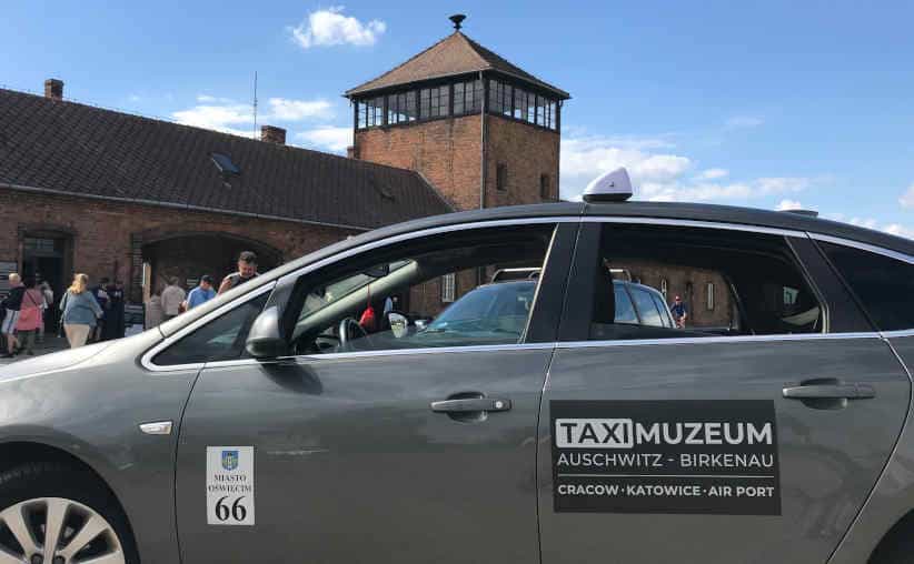 A sliver cab with TAXI MUZEUM written on the side door, in front of Birkenau main gate