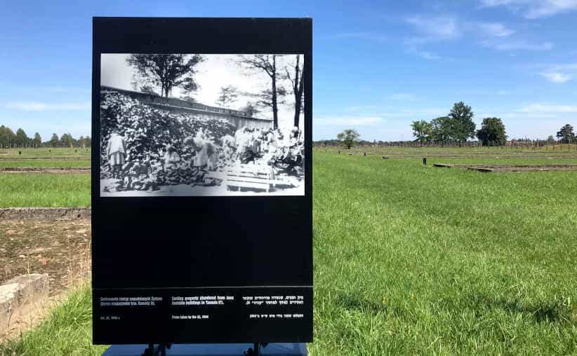A board with a photo of prisoners sorting shoes in Canada with the barracks' ruins in the background at Auschwitz-II-Birkenau.