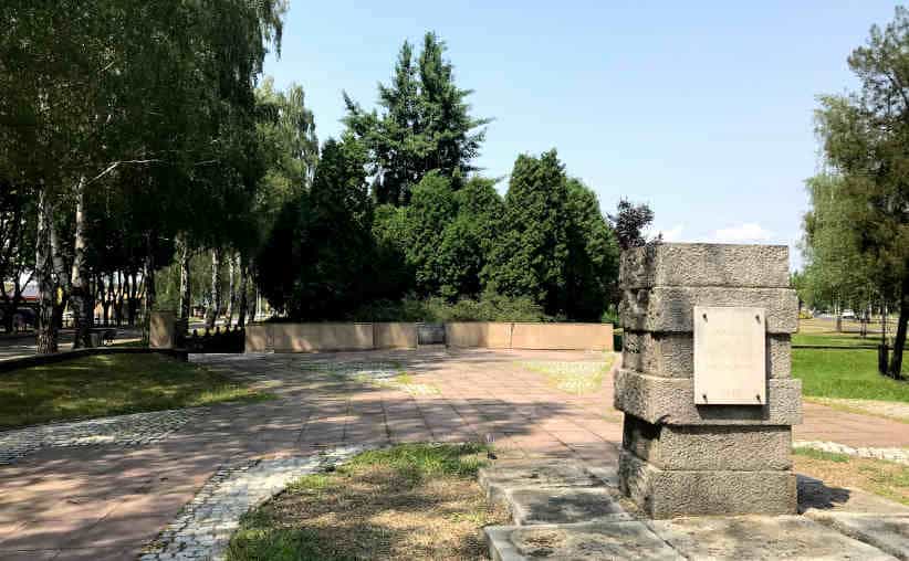 An info board on a stone statue, with tiles on the ground and trees around at the Grave Of The Last Victims Of Auschwitz.